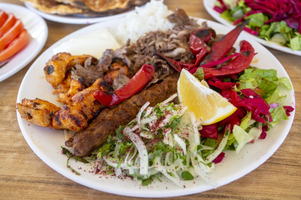 Plato blanco con brochetas de kebab de carne, trozos de pollo especiado, arroz, rodaja de limón y ensalada fresca de lechuga, cebolla y repollo.