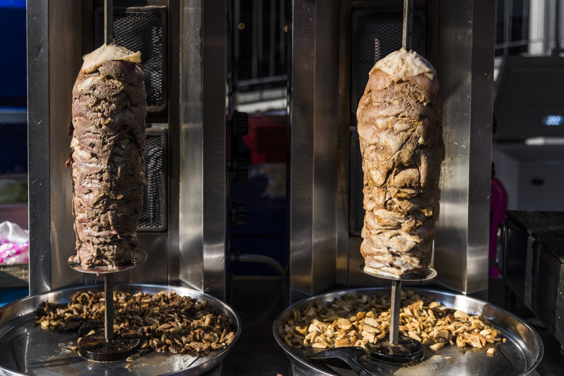 Dos trompos verticales con capas de carne asándose, sobre brasas, con bandejas metálicas recogiendo la carne cortada.