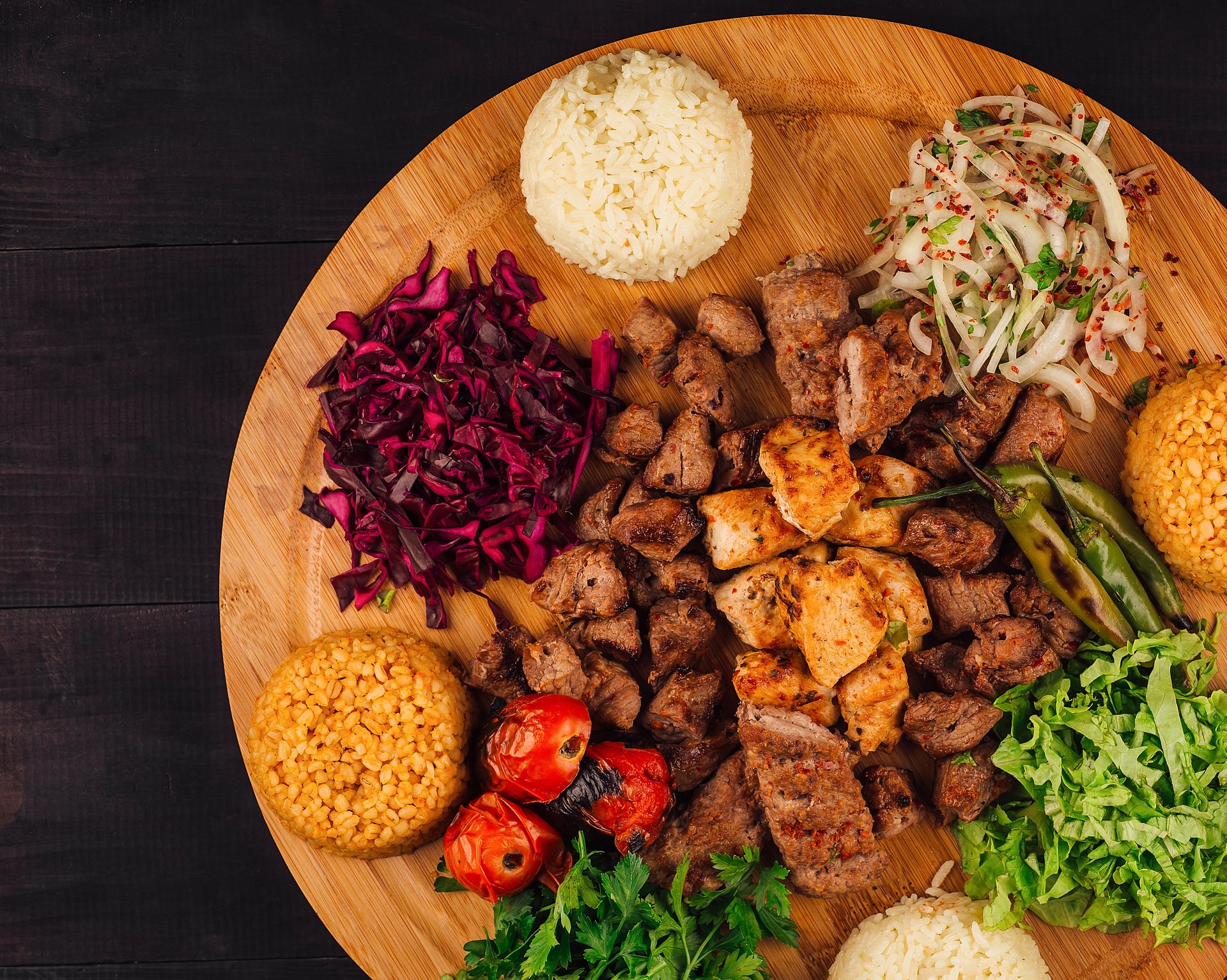 Vista aérea de una tabla de madera con varios tipos de carnes asadas, arroz, bulgur, ensaladas de repollo y lechuga, tomates asados y hierbas frescas.