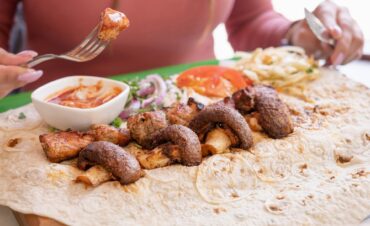 Brochetas de carne y champiñones servidas sobre una lámina de pan plano, acompañadas de salsa en cuenco y guarnición de ensalada.